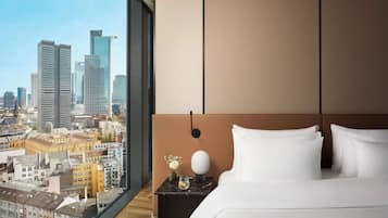 Ein Hotelzimmer mit großem Fenster und Blick auf eine Stadtlandschaft, ein Bett mit weißen Leinen, ein Nachttisch mit Blumen in einer Vase und ein modernes Kopfstück.