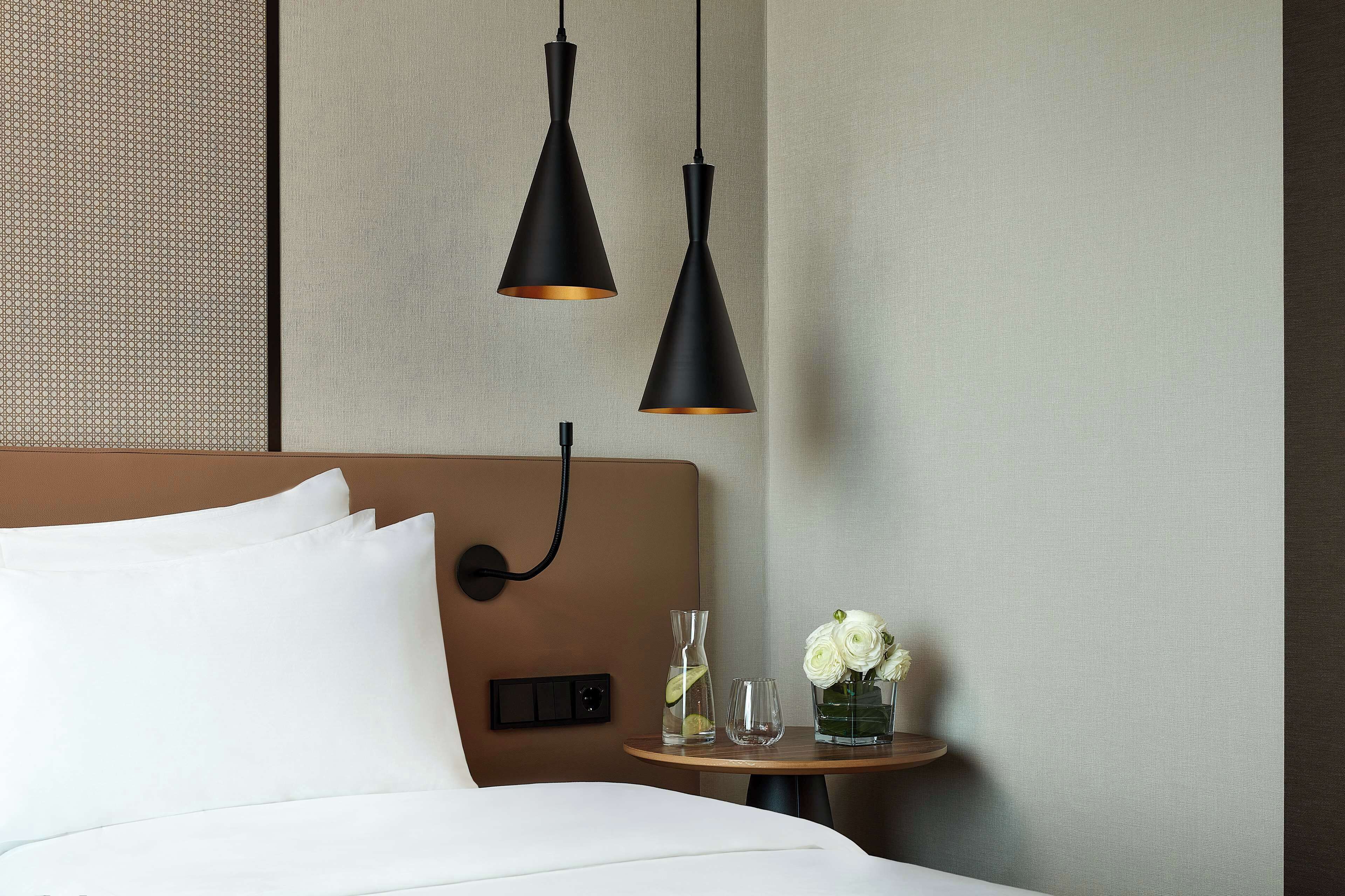 Ein Hotelzimmer mit einem Bett, zwei Pendelleuchten, einem Nachttisch mit Blumenvase und einer strukturierten Wand.