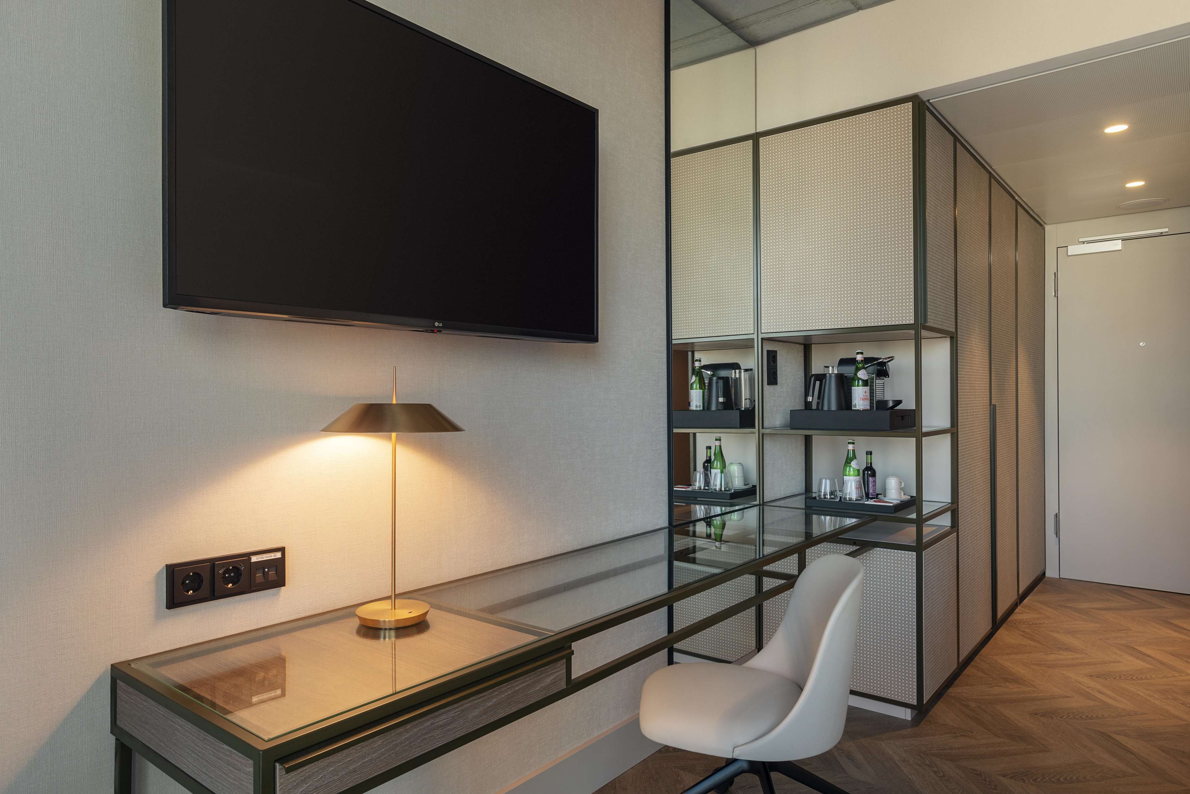 Una habitación moderna con una televisión de pantalla plana montada en la pared, un escritorio con lámpara, una silla y un panel de vidrio con estantes que contienen botellas y copas.