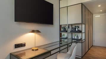 A modern room with a flat-screen TV mounted on the wall, a desk with a lamp, a chair, and a glass partition with shelves containing bottles and glasses.