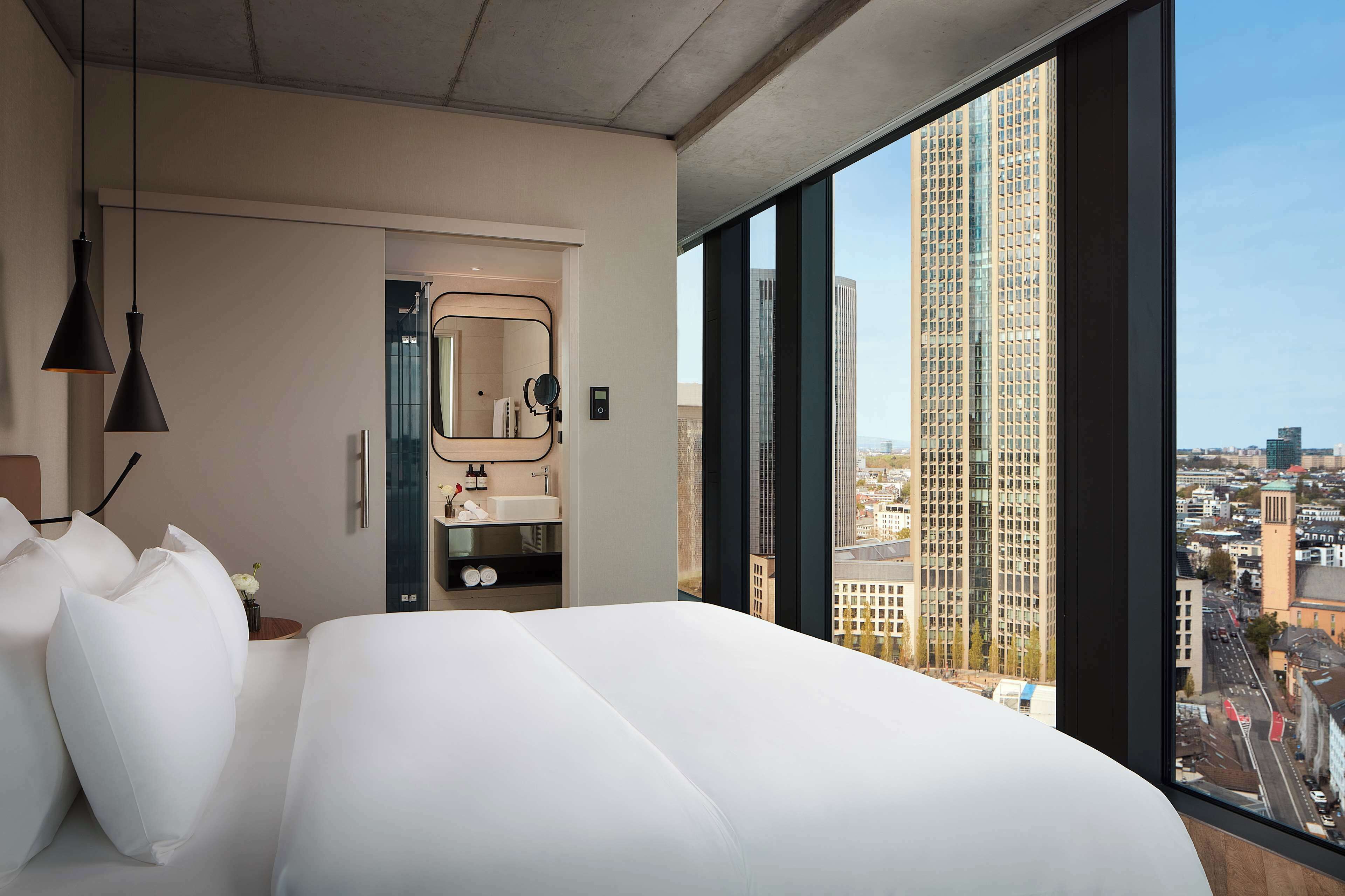 Una habitación de hotel moderna con una cama grande, un baño con espejo y lavamanos, y vistas a la ciudad a través de amplios ventanales.
