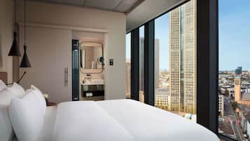 A modern hotel room with a large bed, a bathroom with a mirror and sink, and a city view through large windows.