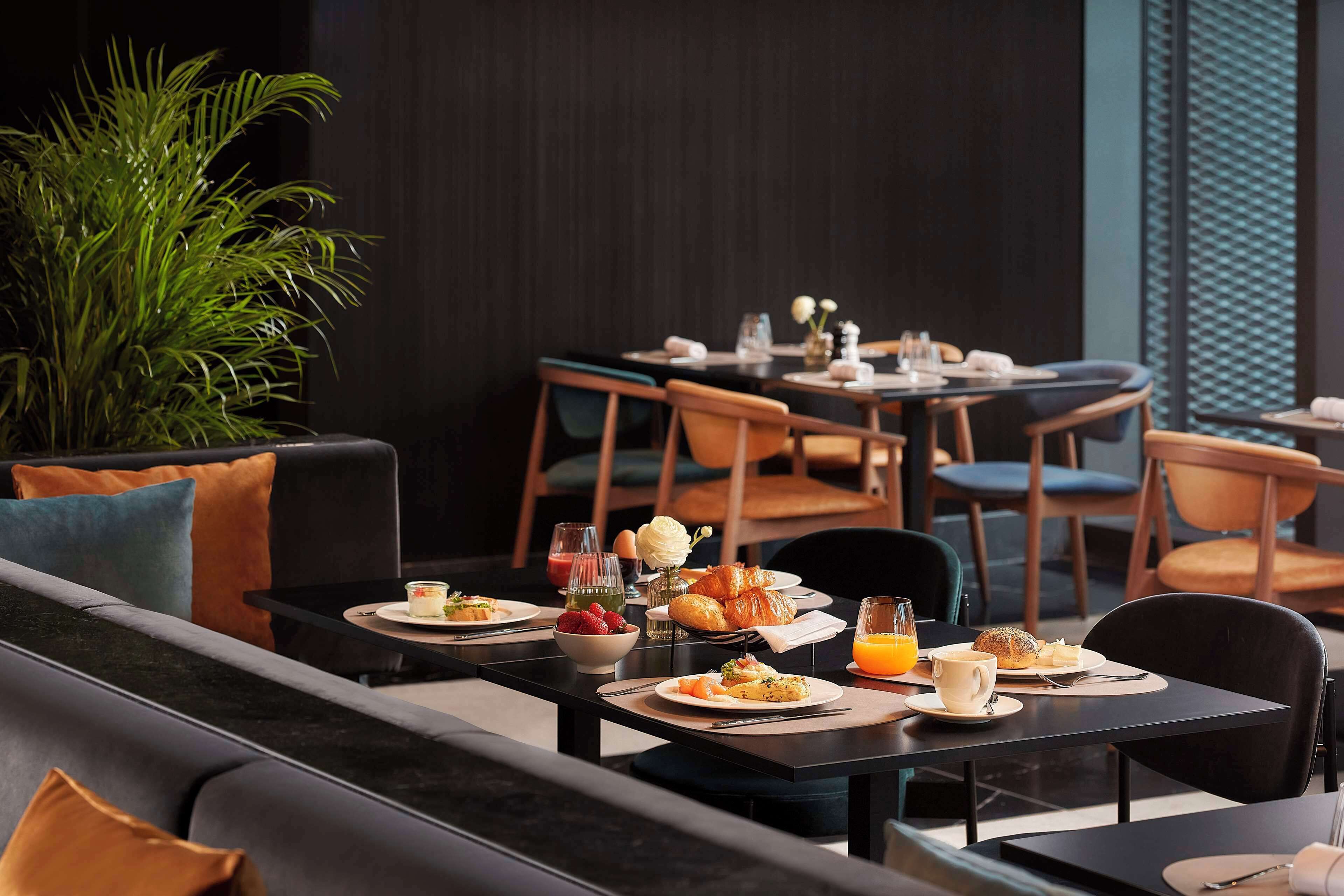 Un comedor moderno con mesas y sillas negras, diversas opciones para el desayuno y una planta verde grande.