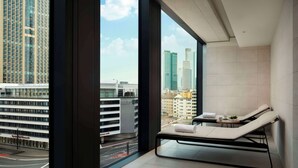 A modern hotel room with a large window offering a city view, a sun lounger, and a small table with a towel and a fruit bowl.