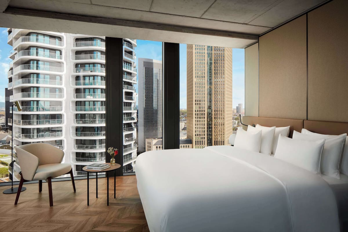 Een moderne hotelkamer met een groot bed, een stoel, een klein tafeltje en uitzicht op hoge gebouwen.