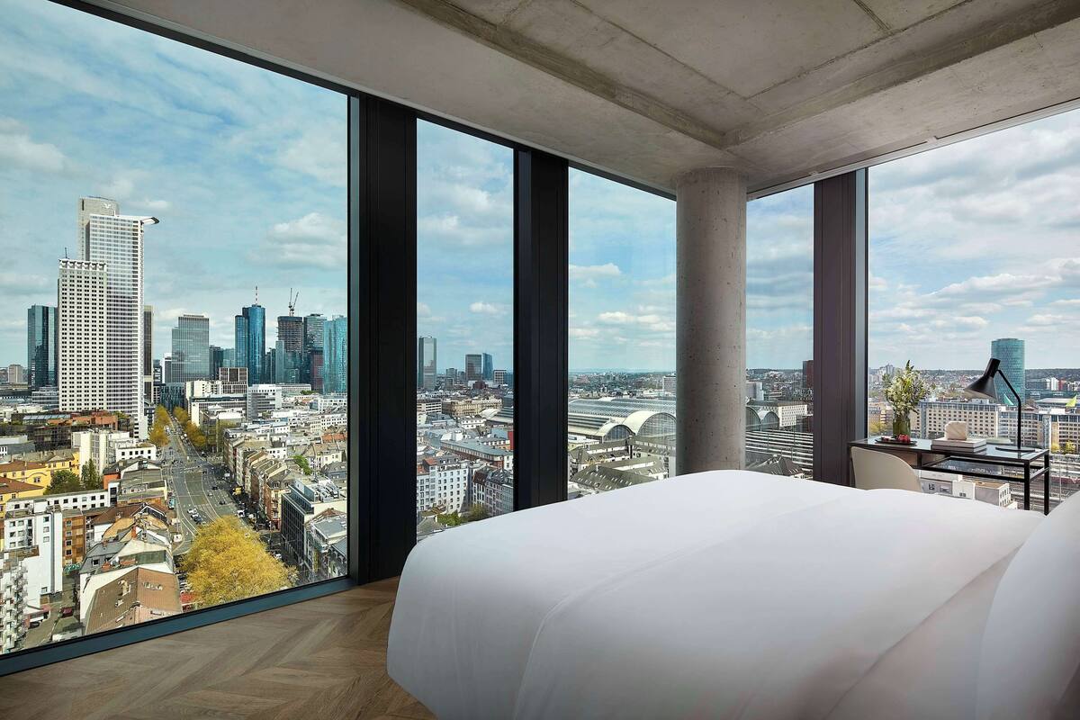 Hotelkamer met een groot bed, een bureau en uitzicht op een stadsgezicht door grote ramen.