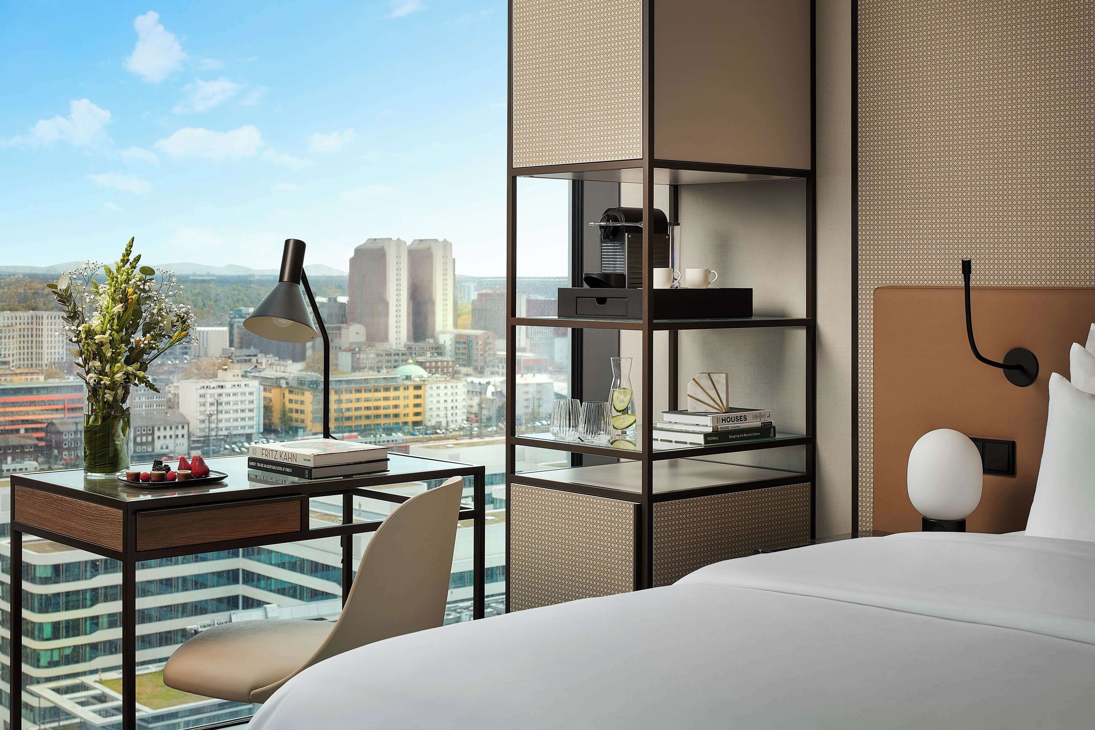 Et moderne hotelværelse med udsigt over byen, et skrivebord med lampe, en bogreol og en seng med sengegavl.