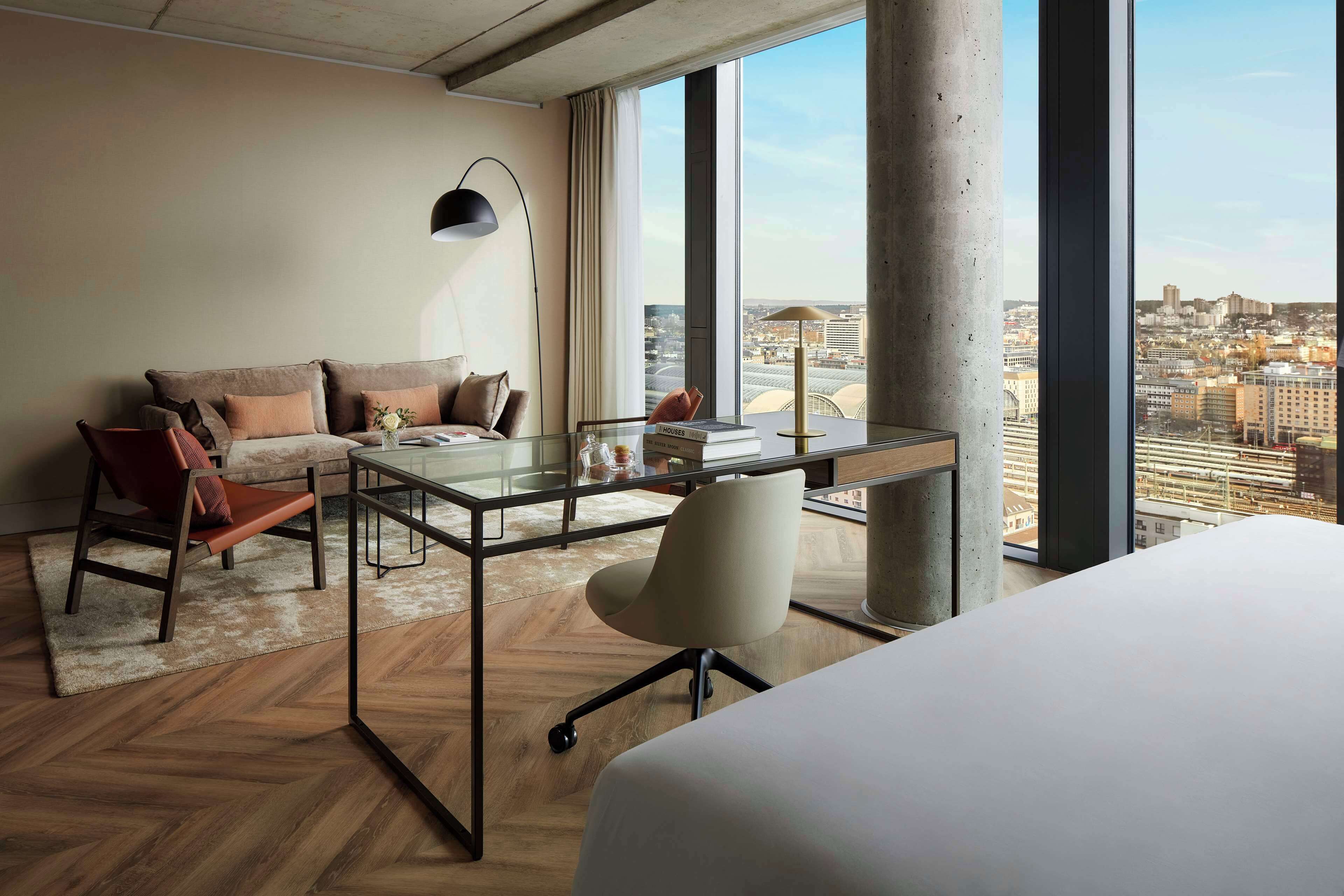 A modern hotel room with a large window, a desk, a chair, a sofa, and a bed.