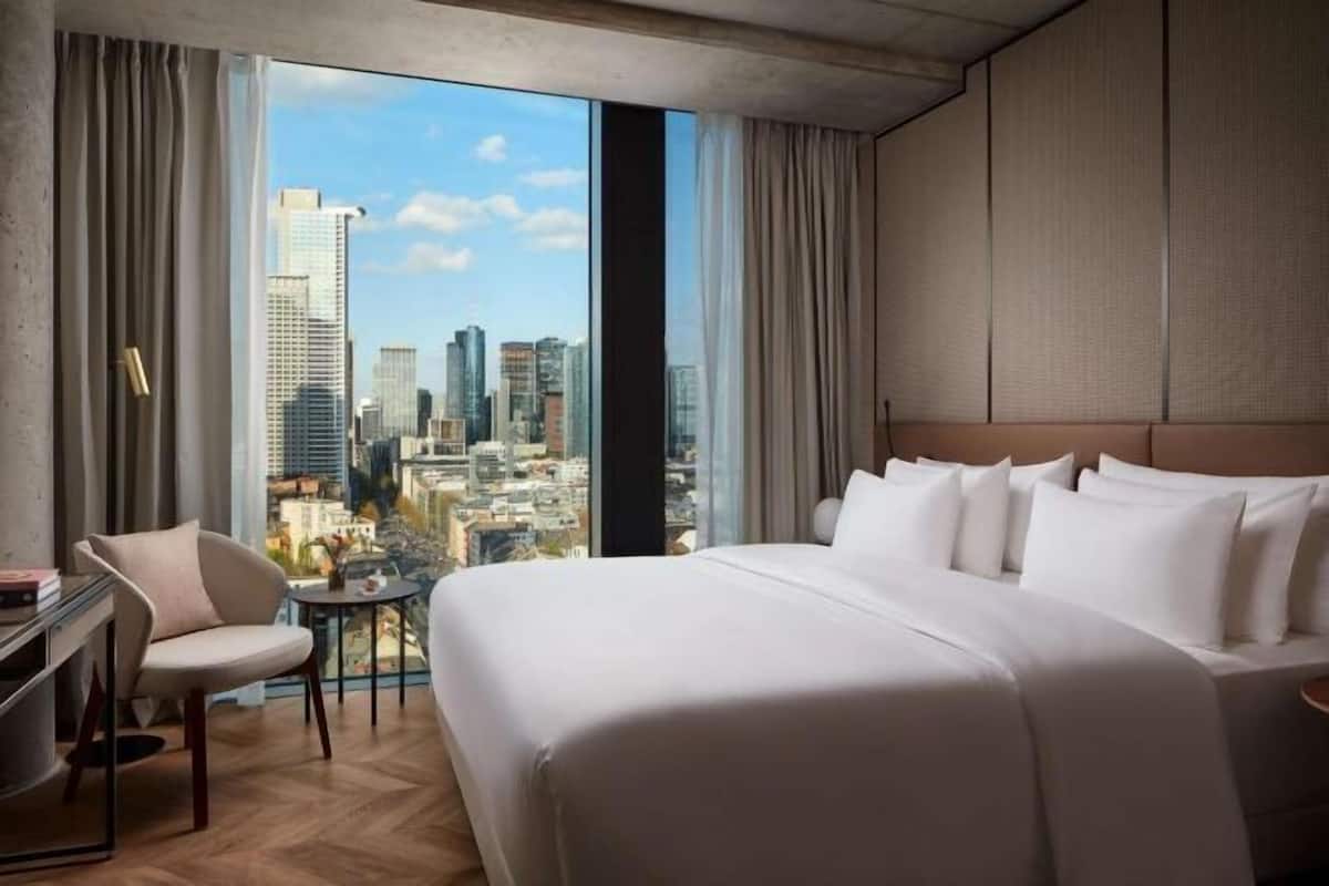 Een hotelkamer met een groot bed, een bureau en een stoel, met uitzicht over de stad door grote ramen.