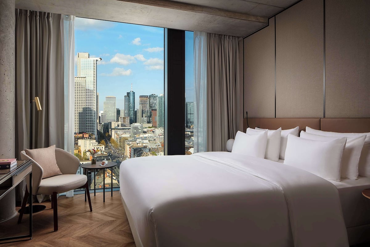 Een hotelkamer met een groot bed, een stoel, een klein tafeltje en een stadszicht door grote ramen.