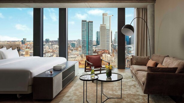 A modern hotel room with a large bed, a sofa, a small table, and a view of the cityscape through large windows.