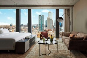 A modern hotel room with a large bed, a sofa, a small table, and a view of the cityscape through large windows. - NH Collection Frankfurt Spin Tower (Frankfurt)