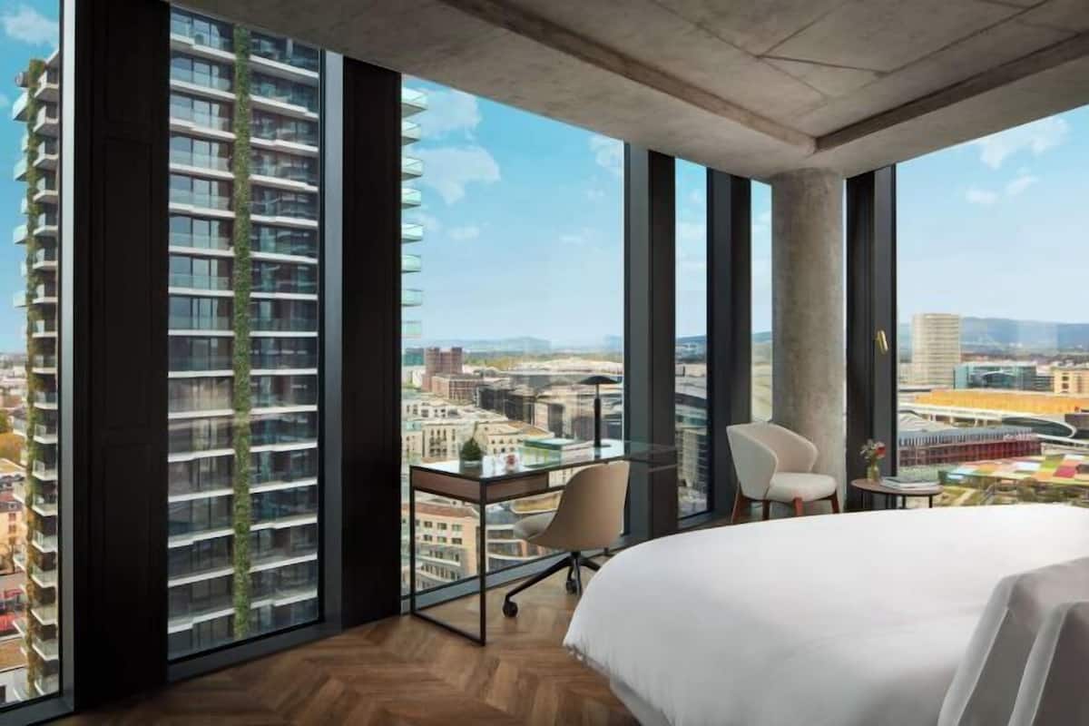 Een moderne hotelkamer met een groot bed, een bureau en een stoel. De kamer heeft grote ramen met uitzicht op een stadsbeeld en een hoog gebouw.