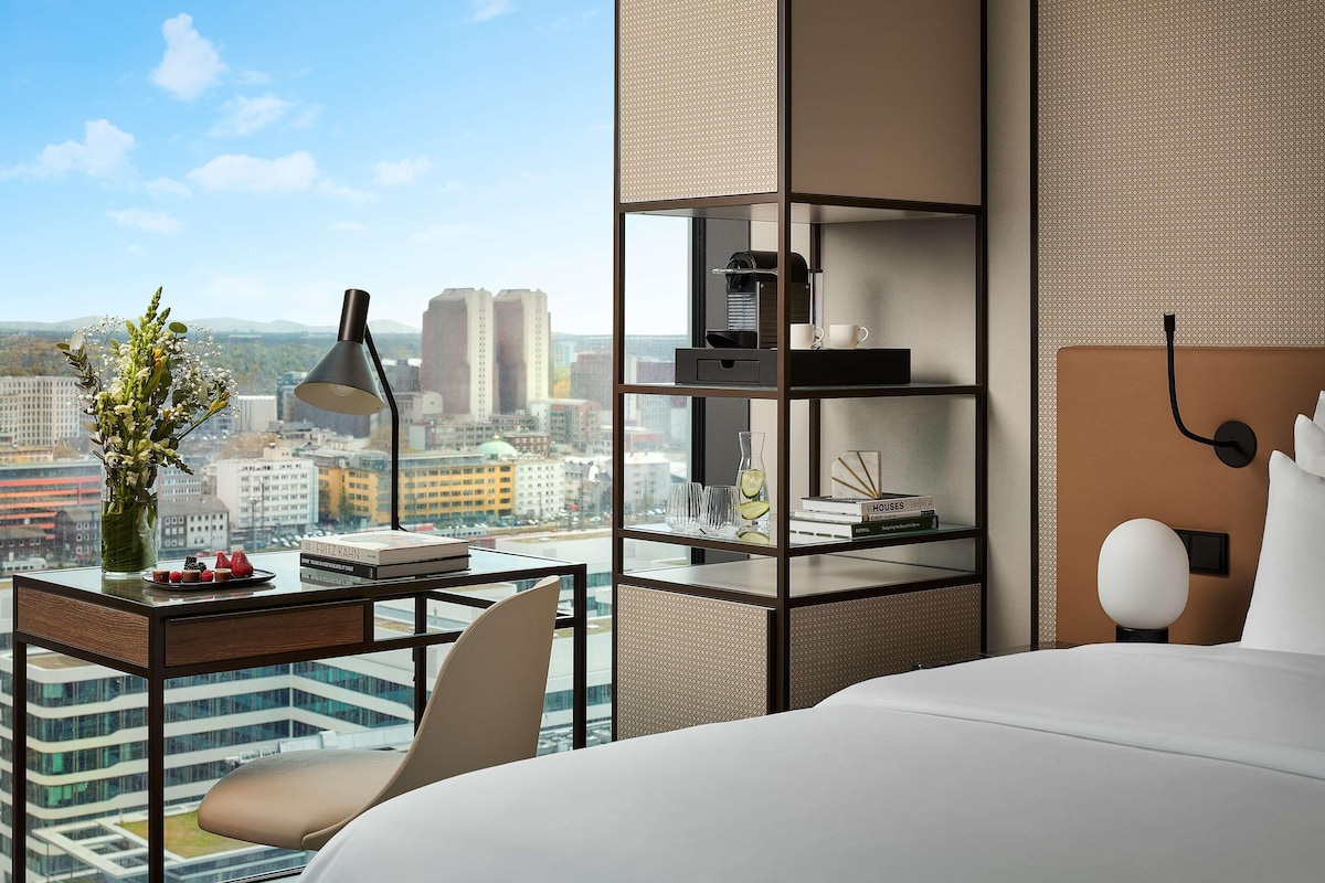 A modern hotel room with a city view, a desk with a lamp, a coffee machine, and a bookshelf.