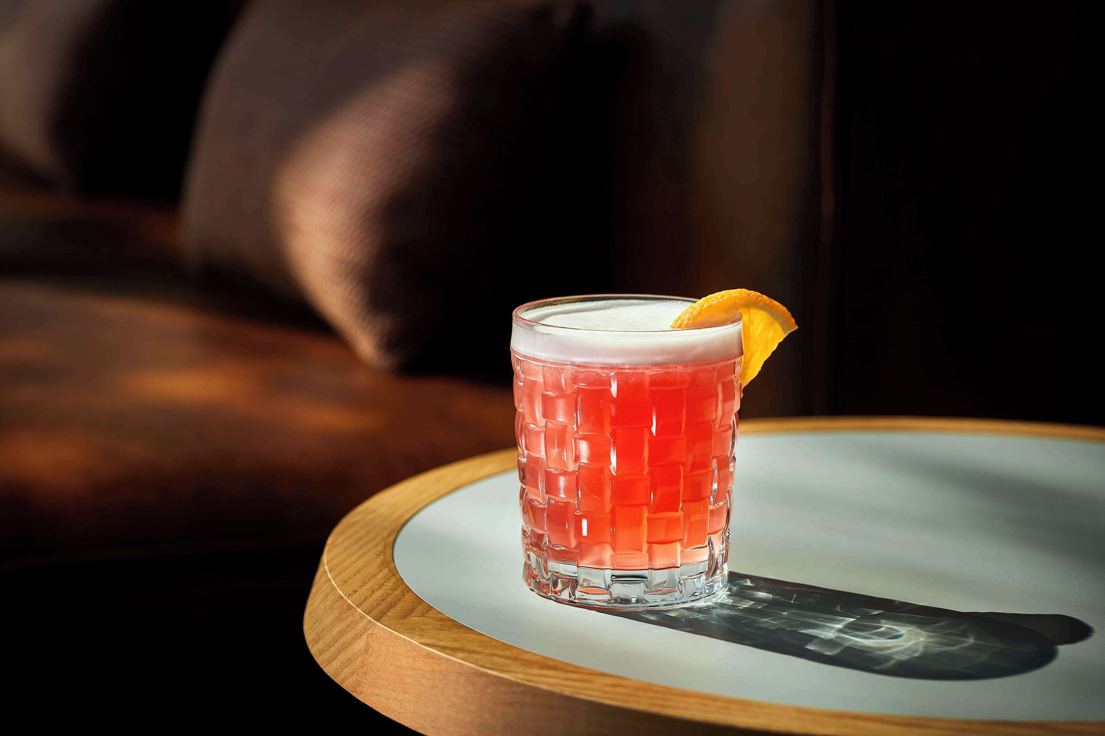 Um copo de cocktail com uma bebida vermelha e um enfeite de laranja sobre uma mesa de madeira redonda.