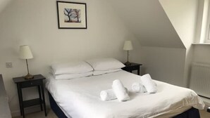 Een net opgemaakt bed met witte lakens en handdoeken, twee nachtkastjes met lampen, een ingelijst foto aan de muur en een radiator.