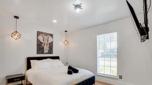 A bedroom with a bed, a nightstand, a wall-mounted TV, and a window with blinds.