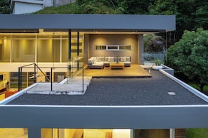 Ein modernes Haus mit Glasfassade, einer Holzterrasse und einer Sitzecke inklusive Sofa und Couchtisch.