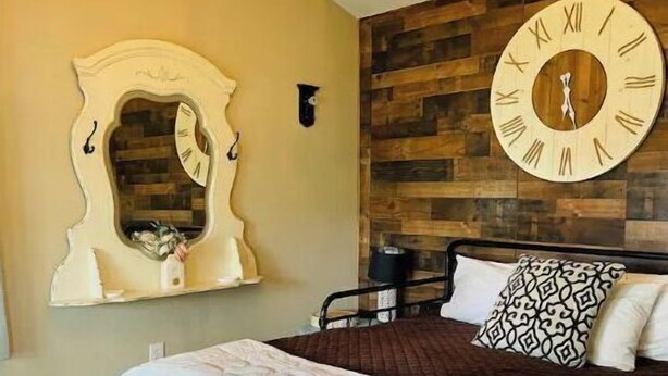 A bedroom with a large wooden headboard, a round wall clock, a chandelier, a mirror, and a bed with multiple pillows. - Cozy, 1 bedroom Cottage with Mountain views! (Tolleson)