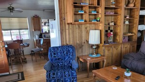 A cozy living room with a blue patterned armchair, a wooden coffee table, and a bookshelf filled with decorative items.