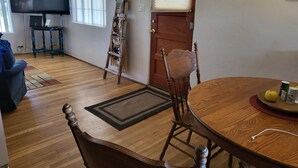 A living room with a wooden floor, a TV, a dining table with chairs, a clock on the wall, and a door with a window.