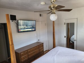Ein Schlafzimmer mit einem Bett, einem hölzernen Schminktisch, einem Deckenventilator und einem an der Wand montierten Fernseher, der eine Nachrichtensendung zeigt.