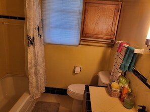 Ein Badezimmer mit Badewanne, Toilette und Waschbecken. Ein Duschvorhang, ein Fenster mit Jalousien und ein Holzschrank über dem Waschbecken sind vorhanden.