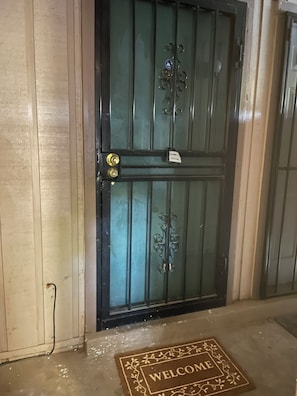 Une porte fermée avec un portail en métal, un paillasson sur lequel est écrit « BIENVENUE », et une prise électrique visible sur le mur.