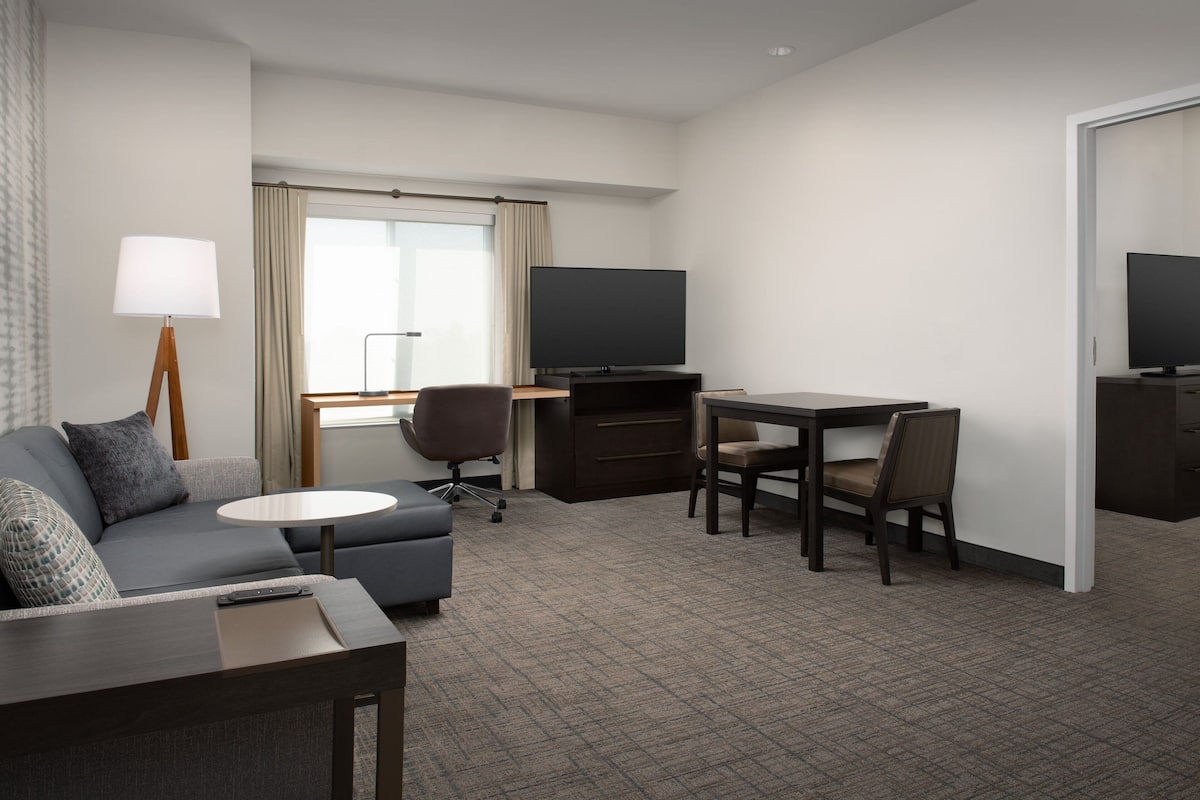 A hotel room with a sofa, a small round table, a desk with a chair, a TV, and a large window with curtains.