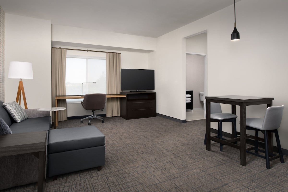 A hotel room with a sofa, a desk with a TV, a dining table with chairs, and a lamp.