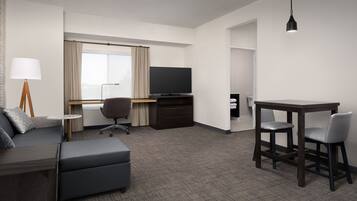 Quarto de hotel com sofá, escrivaninha com televisão, mesa de jantar com cadeiras e luminária.
