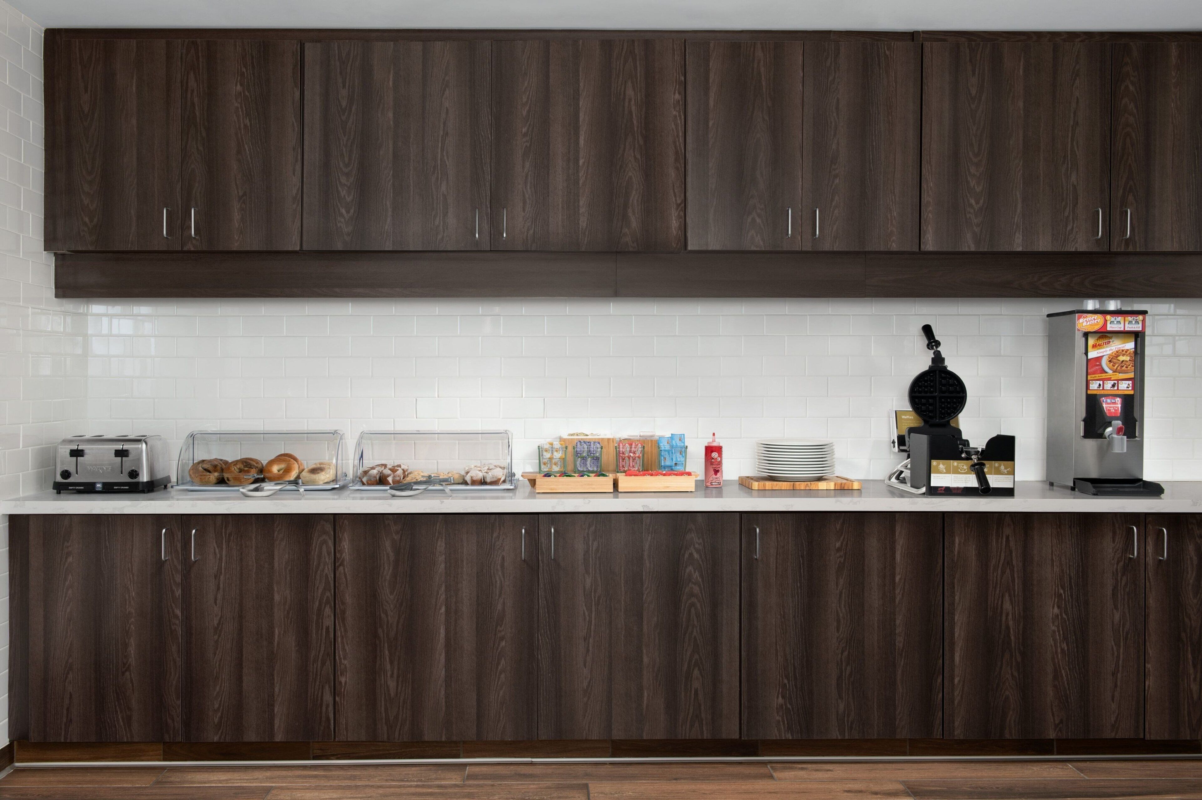 Uma cozinha com armários de madeira escura, painel de azulejos branco, torradeira, máquina de waffles e cafeteira.