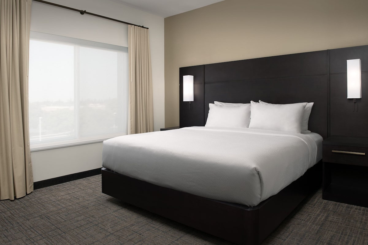 A hotel room with a large bed, a window with curtains, a dark headboard, and bedside lamps.