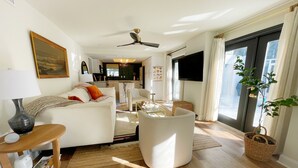 Un salon moderne avec un canapé blanc, une table basse et un téléviseur. On y trouve également un ventilateur de plafond, une grande fenêtre avec des rideaux et une plante.