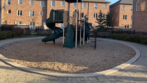 A playground with a slide and climbing structures, surrounded by a gravel area and a brick building in the background.