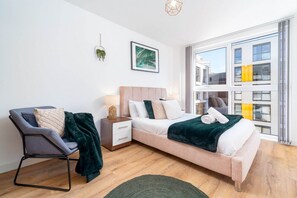 Ein modernes Schlafzimmer mit Bett, Sessel, Nachttisch, Fenster und einer Wandpflanze.