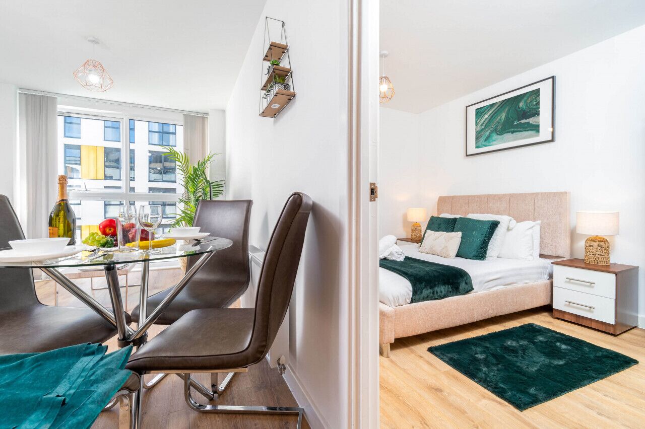 Une chambre d’hôtel moderne avec une table à manger en verre, des fauteuils en cuir, un lit agrémenté d’un plaid vert et une vue sur la ville grâce à de grandes fenêtres.