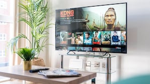 Una sala de estar moderna con un televisor de pantalla plana montado sobre una mesa de vidrio, una planta en maceta y un control remoto sobre la mesa.