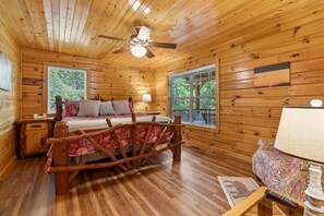 Ein Schlafzimmer einer Holzhütte mit einem Bett, zwei Fenstern, einem Deckenventilator und einem Nachttisch.