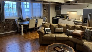 A living room with a brown leather sofa, a glass coffee table, and a dining area with a wooden table and chairs. A kitchen with white cabinets and a microwave is visible in the background. - Historic High School Loft 4 min drive from Oaklawn! (Hot Springs)