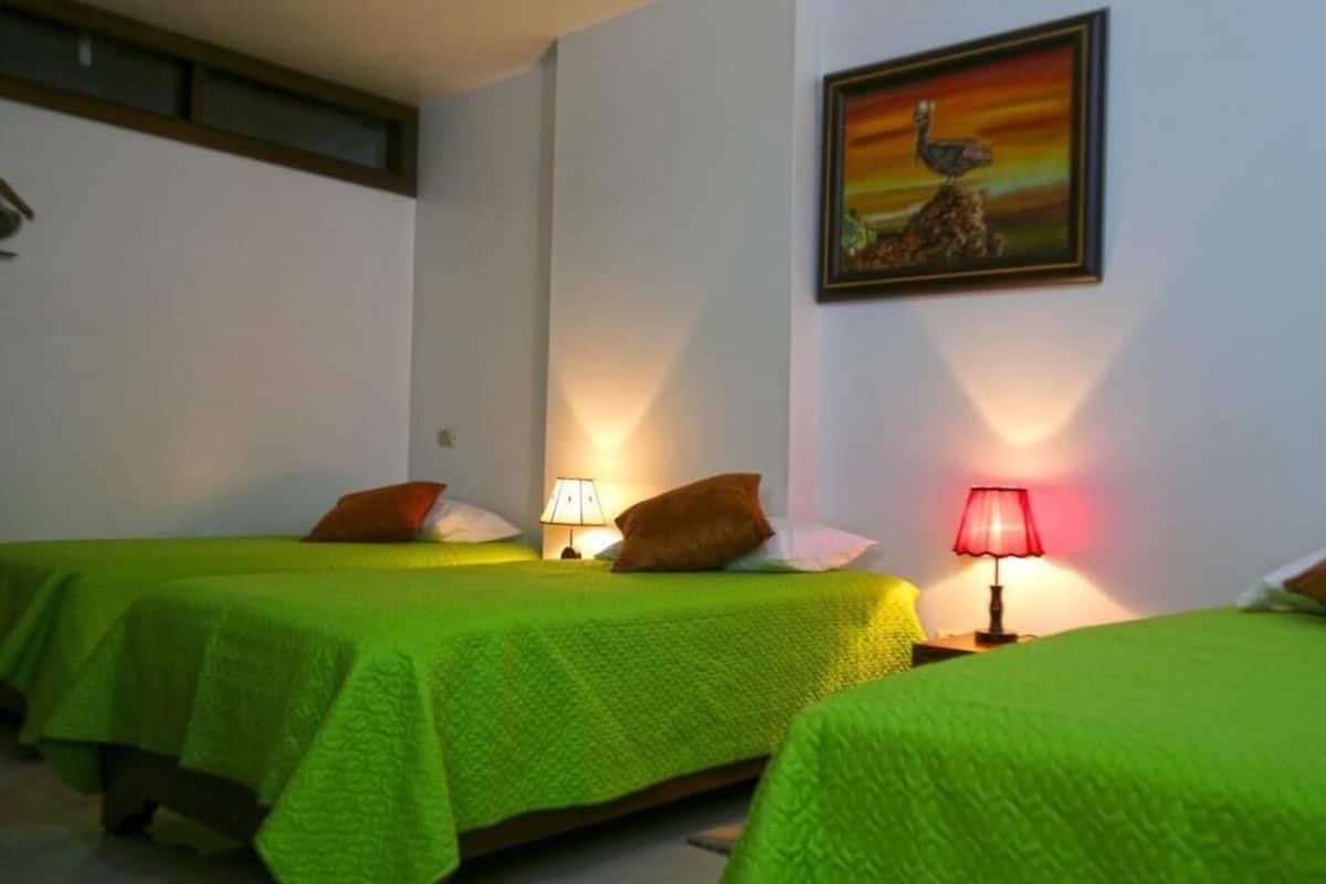 A hotel room with two single beds, each with a green bedspread and a pillow. There is a painting on the wall above the beds.