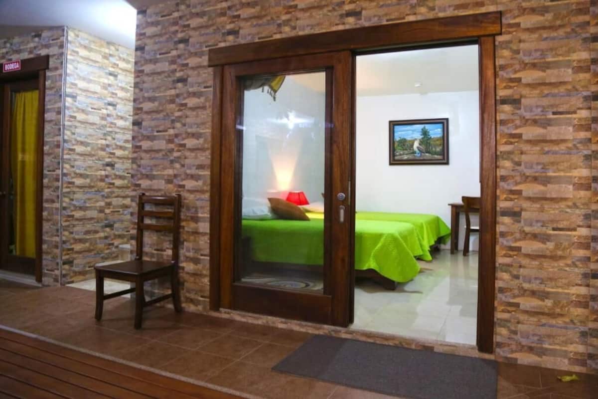 A room with a sliding glass door leading to a bed with green bedding, a wooden chair, and a framed picture on the wall.