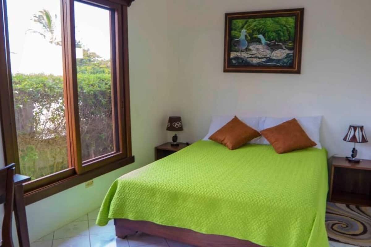 A bedroom with a bed, a window, a chair, and a painting on the wall.