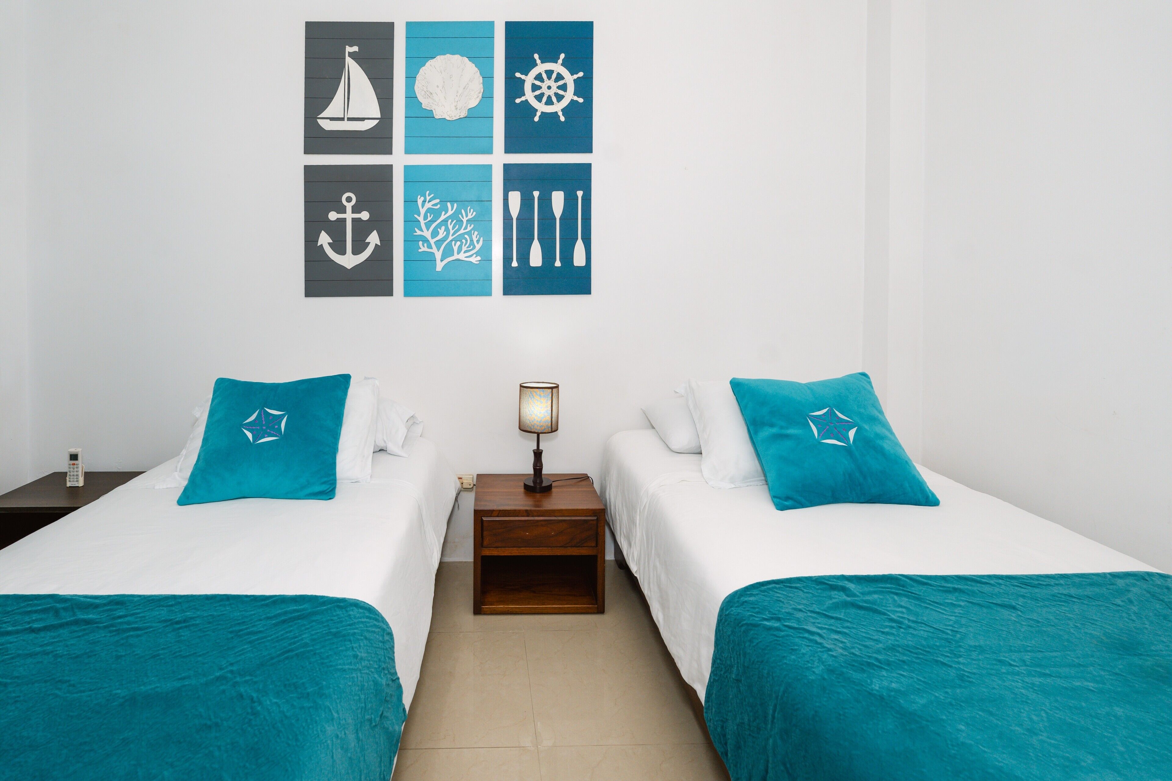 Habitación de hotel con dos camas, cada una con cubrecama color turquesa y almohadas a juego. Hay una mesita de noche de madera pequeña entre las camas con una lámpara encima. Las paredes están decoradas con arte temático náutico.
