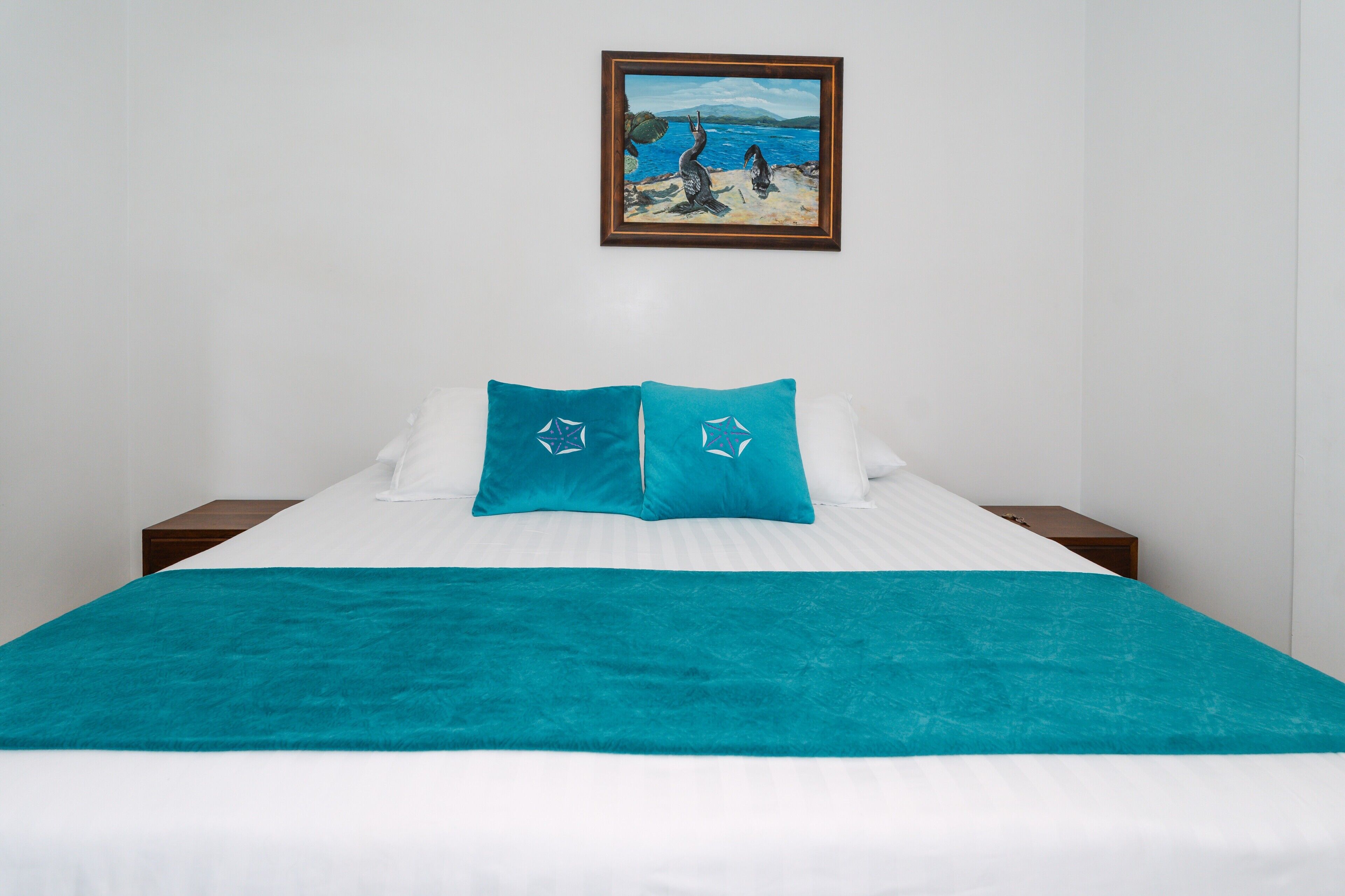 Una cama con colcha y almohadas turquesa, un cuadro enmarcado encima y una pared blanca.