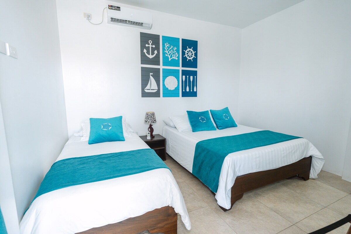 A hotel room with two beds, each with a teal bedspread and matching pillows, a nightstand with a lamp, and a wall with nautical-themed artwork.