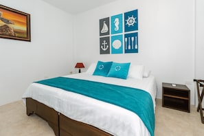 A neatly made bed with a white and teal bedspread, a wooden headboard, and a small bedside table with a lamp. - Starfish Hotel (Puerto Villamil)