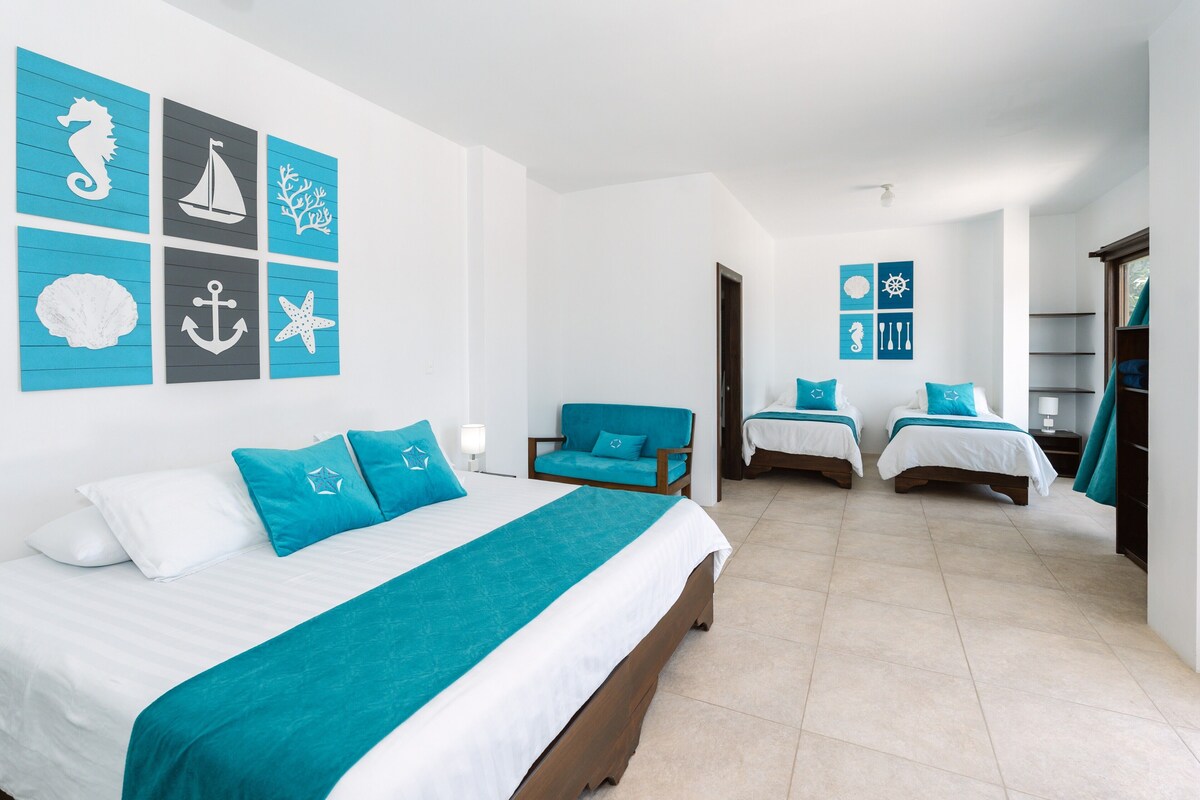 A hotel room with a large bed, two smaller beds, a chair, and a view of the sea.