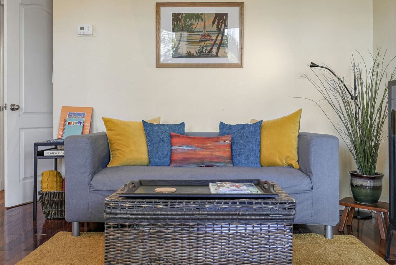 A living room with a grey sofa, yellow and blue cushions, a wicker coffee table, a framed picture on the wall, and a potted plant.