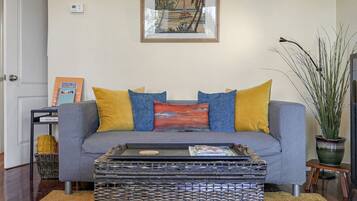 A living room with a grey sofa, yellow and blue cushions, a wicker coffee table, a framed picture on the wall, and a potted plant.